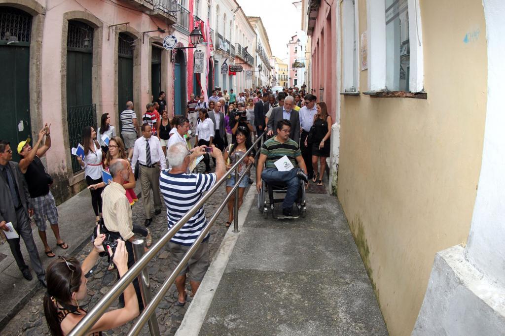 Fotografia da inauguração do Projeto Pelô Acessível. A imagem mostra um homem em cadeira de rodas atravessando a travessia acessível instalada no Pelourinho. O homem usa uma camisa listrada verde e calças jeans. Ao redor da passarela, diversas pessoas observam e fotografam o momento.