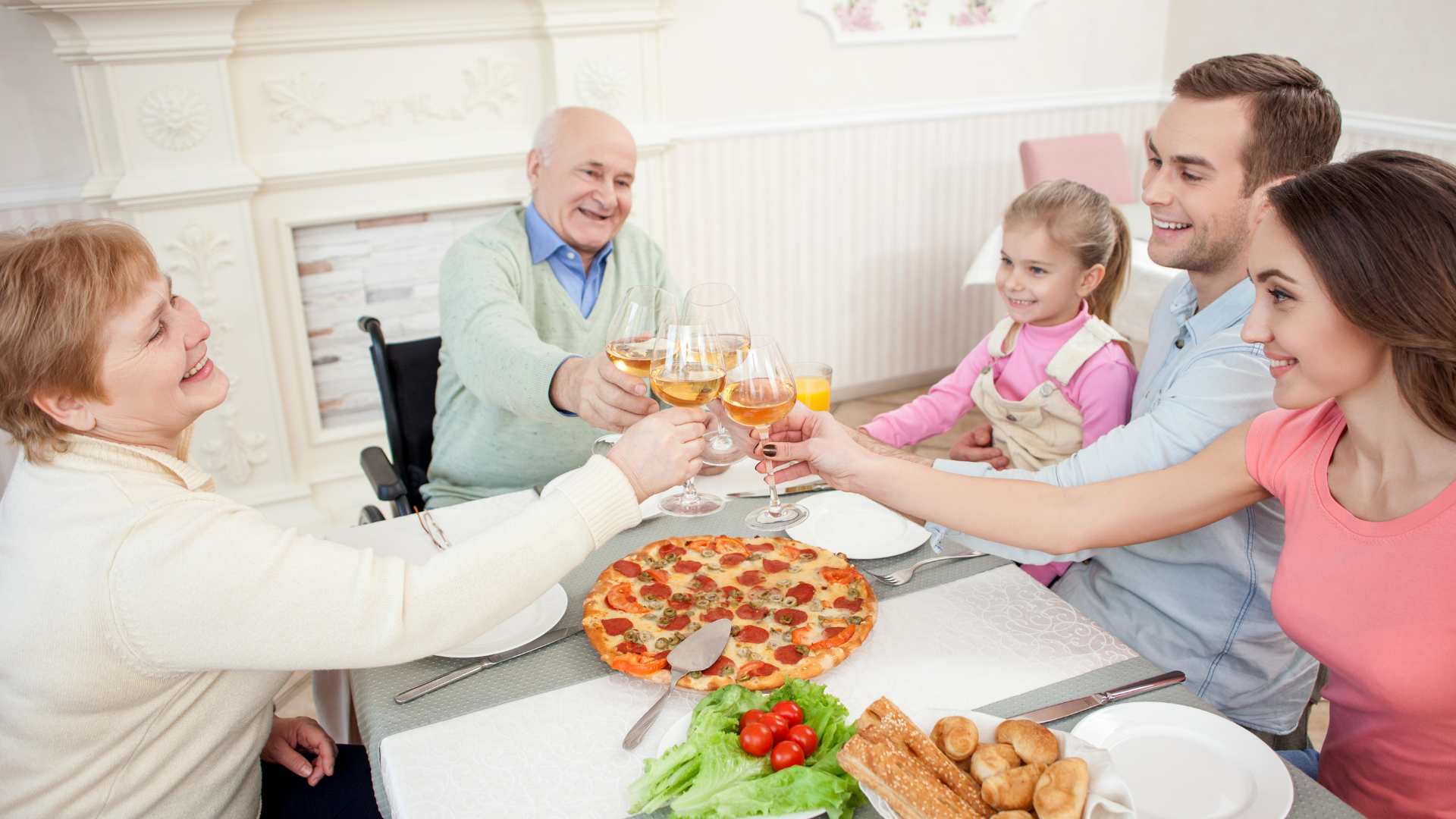 A fotografia mostra pessoas sentadas à mesa, fazendo um brinde. Da esquerda para a direita: uma senhora loira, em torno de 60 à 70 anos, com uma taça de vinho; um senhor grisalho, em torno de 70 anos, em cadeira de rodas, sorrindo e segurando uma taça de vinho; uma menina loira, em torno de 10 anos, sorridente e segurando um copo de suco. A menina está sentada no colo de um homem branco, na faixa dos 30 anos, sorridente, que também segura uma taça. Ao seu lado, uma mulher branca de cabelos castanhos, sorrindo e segurando uma taça. Ao centro do brinde, a mesa possui pratos, talheres e alimentos como pizza, salada e pães.