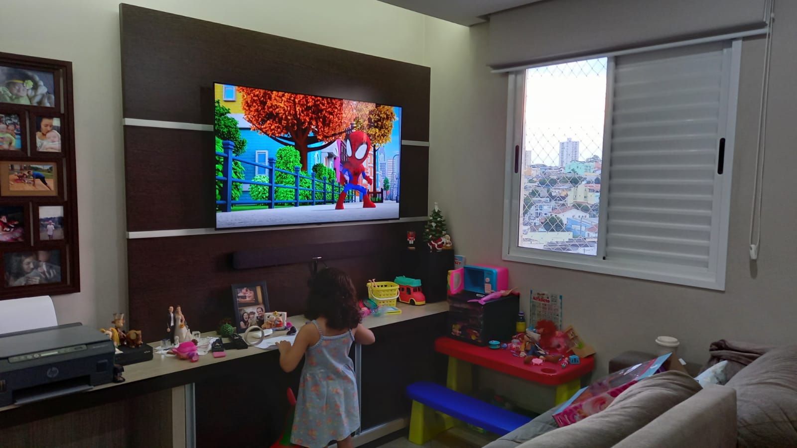Fotografia de sala de televisão, com estante, uma televisão ligada transmitindo desenho infantil, ao seu lado uma mesa de brinquedos e à frente um sofá. Uma menina brinca em frente à televisão.